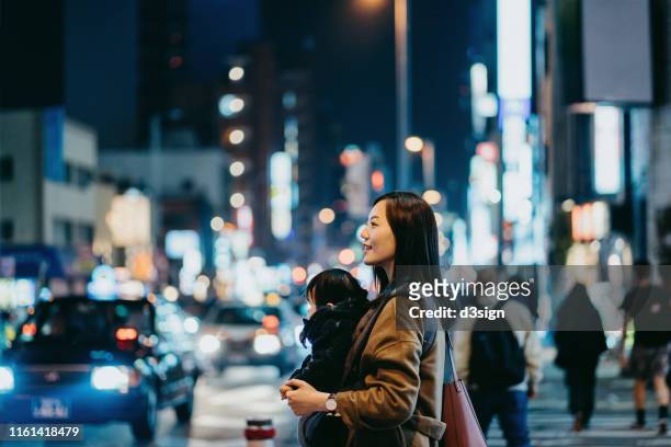 young asian mother crossing street and commuting in busy downtown city with little daughter at night, against crowd of pedestrians and city traffic in the background - mother and daughter on night street stockfoto's en -beelden