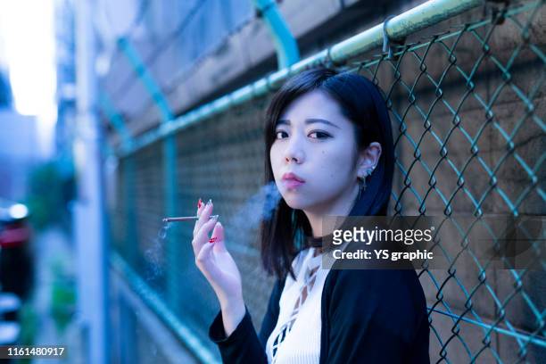 young and beautiful asian woman smoking. she is leaning on a fence. - beautiful women smoking cigarettes stock pictures, royalty-free photos & images
