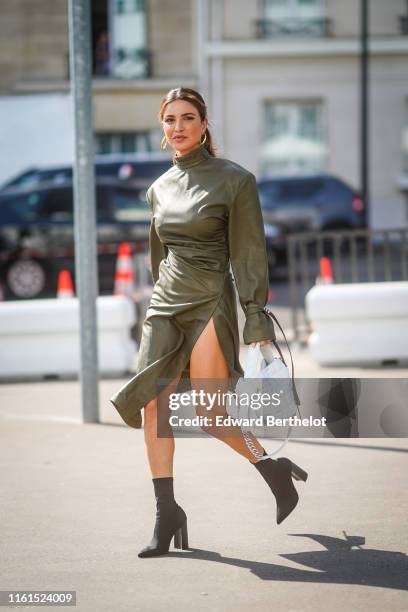 Negin Mirsalehi wears earrings, a khaki leather hi-neck , thigh-slit drape dress, a white quilted Acne Studios bag, black pointy sock-boots, outside...