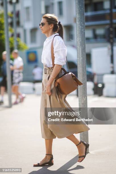 Guest wears sunglasses, a white shirt, a light khaki skirt, a brown Acne Studios bag, black ankle-strap flat sandals, outside Acne, during Paris...