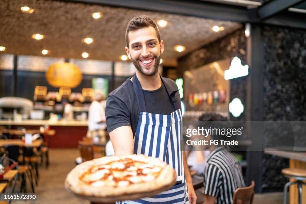 lächelnder kellner, der in die kamera schaut und eine pizza zeigt - waiter serving stock-fotos und bilder