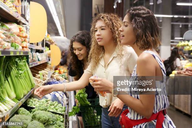 three female latinx millennials shop for vegetables at a grocery store, wearing summer clothes. - latin american and hispanic shopping bags stockfoto's en -beelden