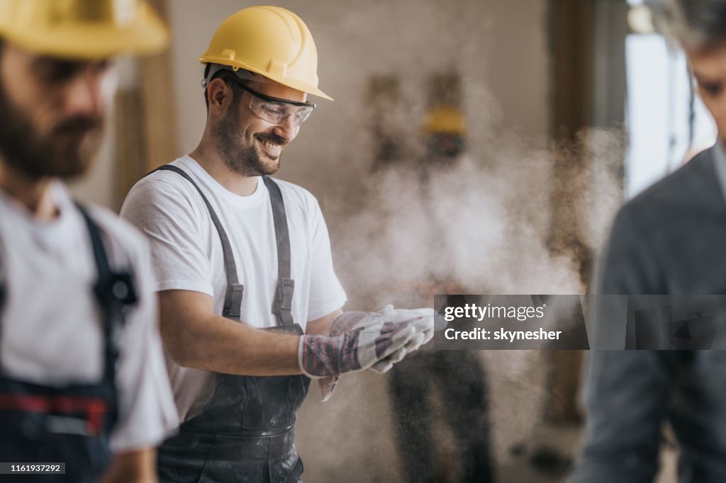 Happy construction worker cleaning his gloves from sawdust at in renovating apartment.