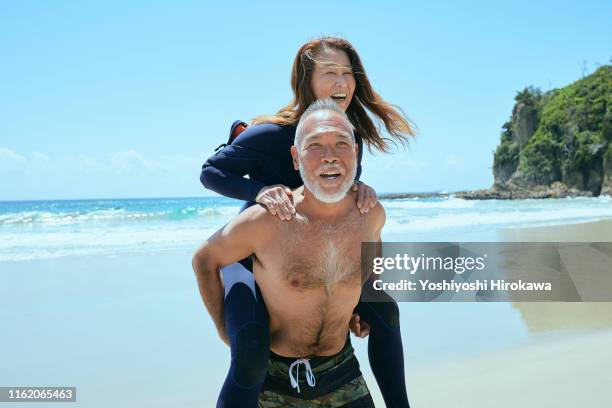 senior man giving wife piggyback ride on beachat beach - japanese couple beach stock-fotos und bilder