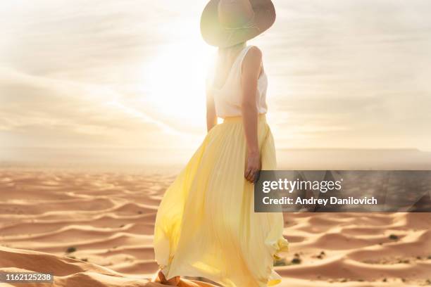 a young caucasian woman in a long skirt, shirt and straw hat standing on top of a sand dune and looks towards the sunrise. sunrise in the merzouga (sahara) desert. - skirt stock pictures, royalty-free photos & images