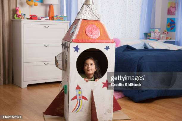 hispanic girl playing inside cardboard rocket - space helmet stock-fotos und bilder