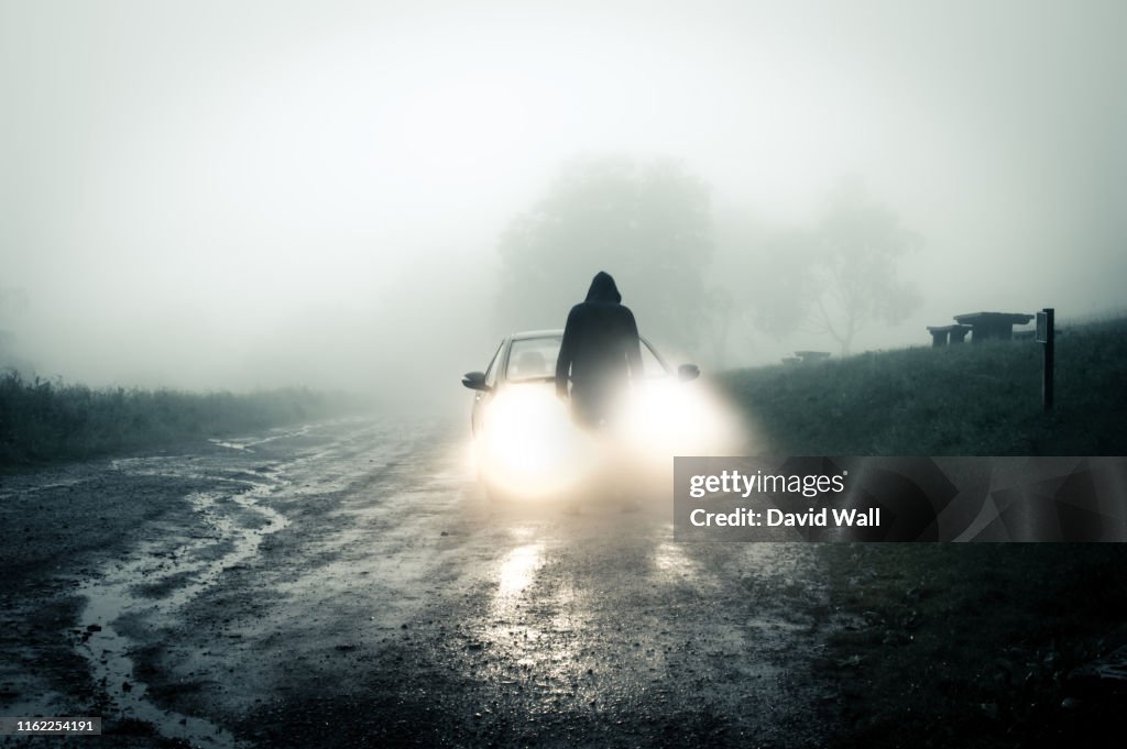 A lone, spooky, hooded figure standing in front of a car looking at an empty misty country road silhouetted at night by car headlights.