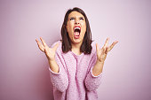 Young beautiful brunette woman wearing a sweater over pink isolated background crazy and mad shouting and yelling with aggressive expression and arms raised. Frustration concept.