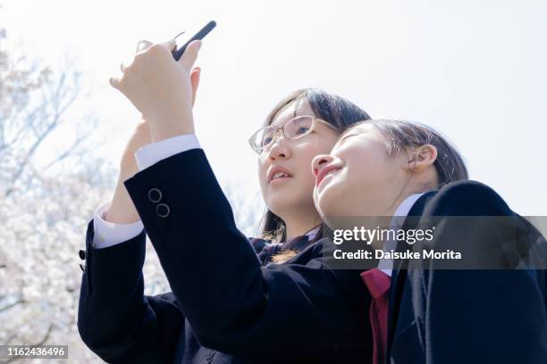 two teenagers in school uniform taking a selfie under cherry blossom trees - female high school student stock pictures, royalty-free photos & images