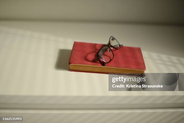 a book and reading glasses on a white shelf in a beautiful sunlight coming through blinds - reading glasses stock pictures, royalty-free photos & images