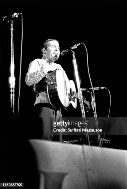 Paul Simon, smiling, right hand on his guitar, with three mikes, speaking to the audience from the stage at the Monterey International Pop Festival,...