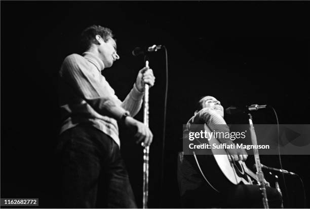 Paul Simon smiling and looking up as Art Garfunkle looks on as he holds a mike stand with two hands, performing on stage at the Monterey...