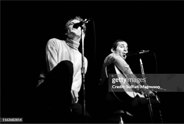 Paul Simon, eyebrows raised and Art Garfunkle with hands clasped in front, right knee in front of his chest, performing on stage at the Monterey...