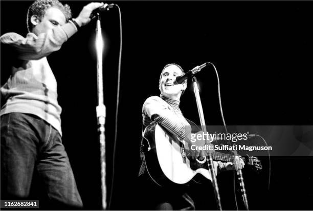 Paul Simon smiling as he sings and plays his guitar and Art Garfunkle, standing with his right hand on a standing microphone, performing on stage at...