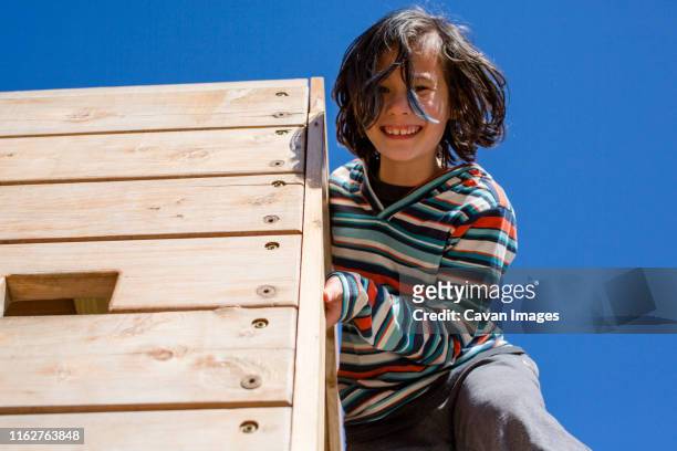 a happy child leans down from a high wooden wall against blue sky - kid looking up to the sky imagens e fotografias de stock
