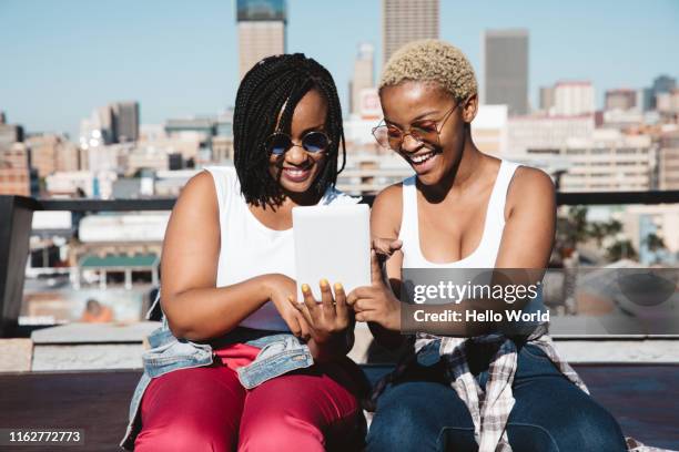 two radiant young women laughing whilst looking at digital tablet - johannesburg stockfoto's en -beelden