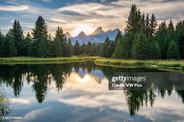 Sunlight creeps behind peaks of Teton Mountain Range with glassy water in the foreground, Grand Tetons National Park, Teton County, Wyoming