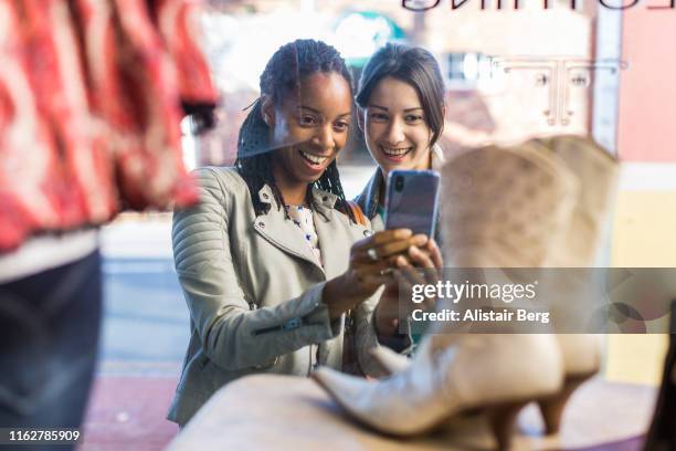 two female friends photographing display in vintage clothes shop window - schaufensterbummel stock-fotos und bilder