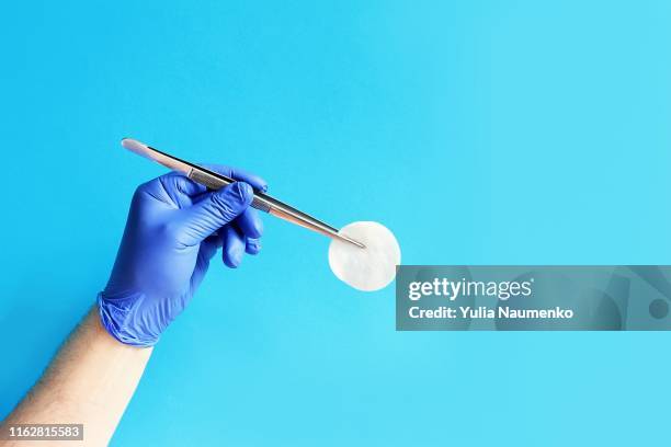 medicine and surgery theme: doctor's hand in a blue glove holding a pair of tweezers with a cotton pad on a blue background, isolated. - pinça utensílio de servir - fotografias e filmes do acervo