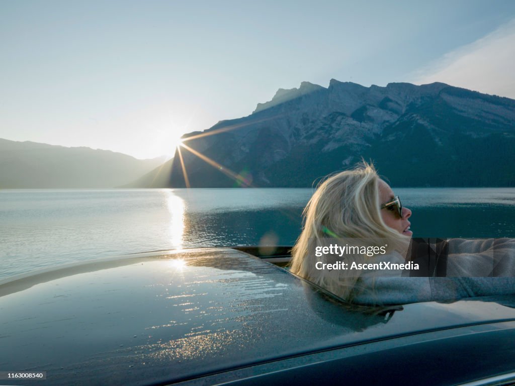 Woman emerges from car sunroof to look at rising sun over the mountains