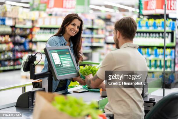 beautiful friendly customer at checkout smiling at male cashier while he scans products in a supermarket - checkout stock pictures, royalty-free photos & images