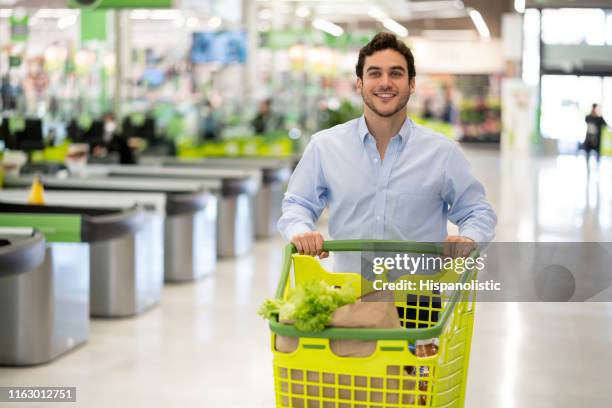 スーパーマーケットで若い独身の男は、カメラの笑顔に直面して購入した食料品でカートを押す - walking away from camera ストックフォトと画像