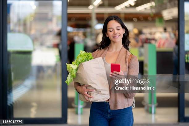 latin american beautiful single woman leaving the supermarket carrying groceries in a paper bag while chatting on smartphone very happy - leaving store stock pictures, royalty-free photos & images