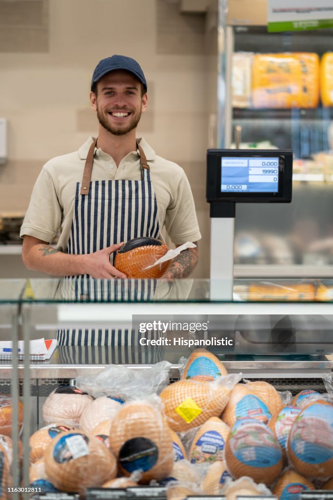 Retrato de vendedor feliz en la sección delicatessen sosteniendo un jamón mientras mira la cámara sonriendo