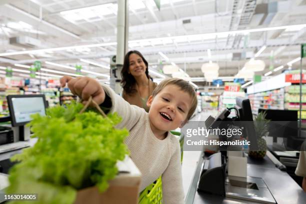 kleine jongen staande in winkelwagen bij de supermarkt overhandigen van een papieren zak met boodschappen en moeder staande op de achtergrond glimlachend bij de camera - retail occupation stockfoto's en -beelden