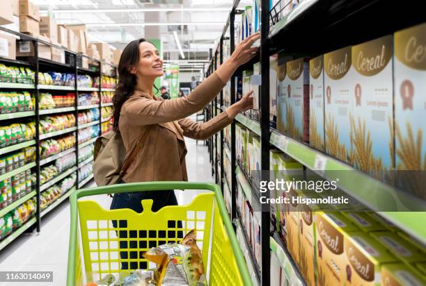 schöne junge frau greift nach einem müsli auf der oberseite regal des supermarktes lächelnd - cereal box stock-fotos und bilder