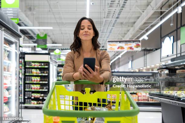 cheerful young beautiful woman checking her shopping list on smartphone while leaning elbows on cart at the supermarket - woman phone market stock pictures, royalty-free photos & images