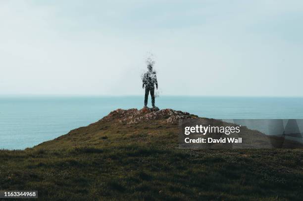 a lone hooded figure standing on top of a hill and disintegrating while looking out across the ocean. - dissolving stockfoto's en -beelden