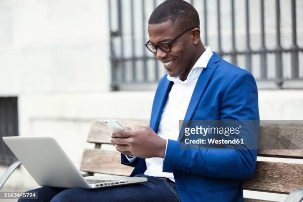 young businessman wearing blue suit jacket sitting on bench and using smartphone - businessman in black suit photos et images de collection