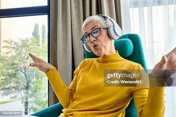 senior woman sitting in armchair, listening music with headphones - vissla bildbanksfoton och bilder