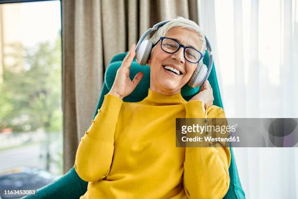 senior woman sitting in armchair, listening music with headphones - 55 59 anni foto e immagini stock