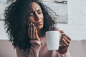unhappy african american woman suffering from toothache and holding coffee cup