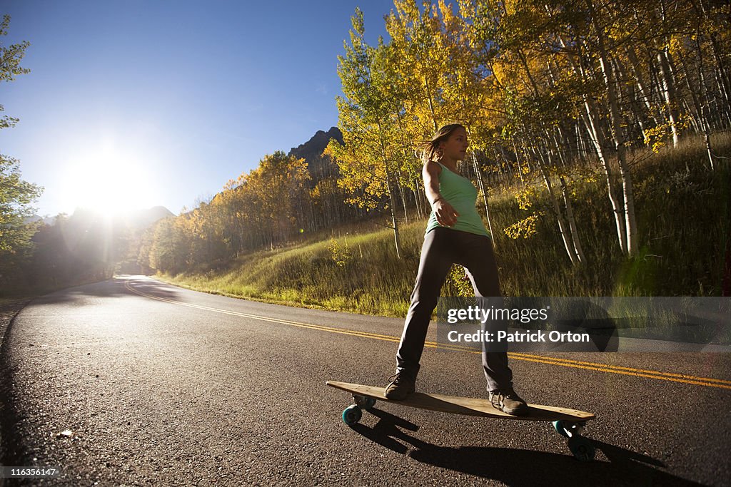 A young woman longboards down a smooth country road through the mountain peaks and gold forests.