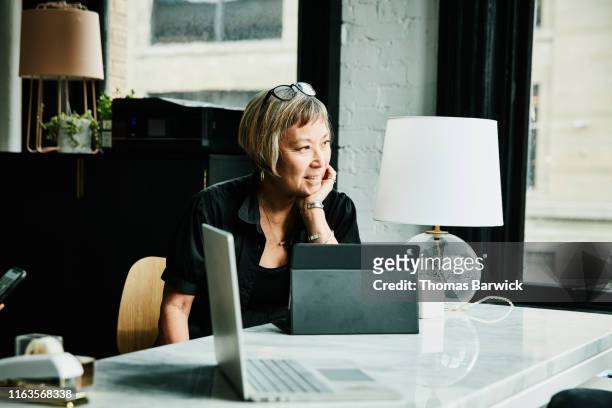 portrait of smiling mature businesswoman seated at desk in creative office - smart windows photos et images de collection