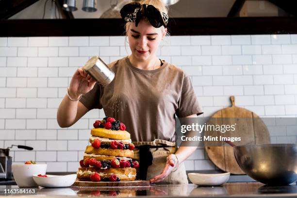 a cook working in a commercial kitchen sprinkling icing sugar over a layered cake with fresh fruit. - woman back stock-fotos und bilder