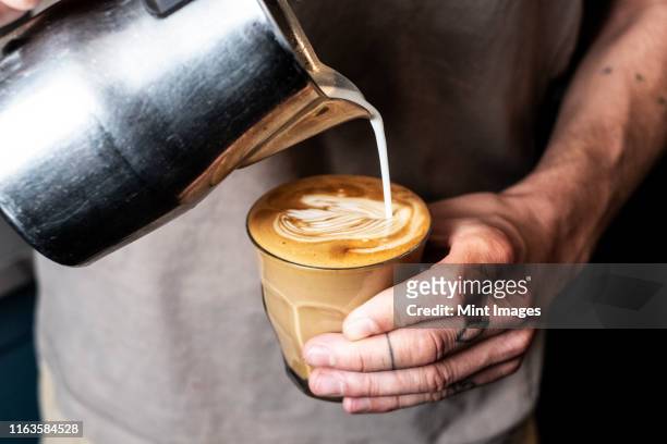 close up of person with tattooed finger pouring milk from metal jug into glass of cafe latte. - fülle stock-fotos und bilder