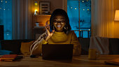 Portrait of Beautiful Smiling Black Girl Sitting at Her Desk Using Laptop to Make a Video Call, Says Hello. In the Evening Girl Talks with Relatives and Friends Using Computer Webcam.