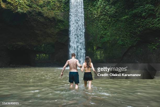 young couple holding hands in river by tibumana waterfall in bali, indonesia - bali waterfall stock pictures, royalty-free photos & images
