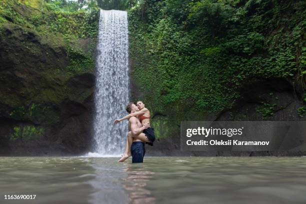 young man carrying young woman in river by tibumana waterfall in bali, indonesia - bali waterfall stock pictures, royalty-free photos & images