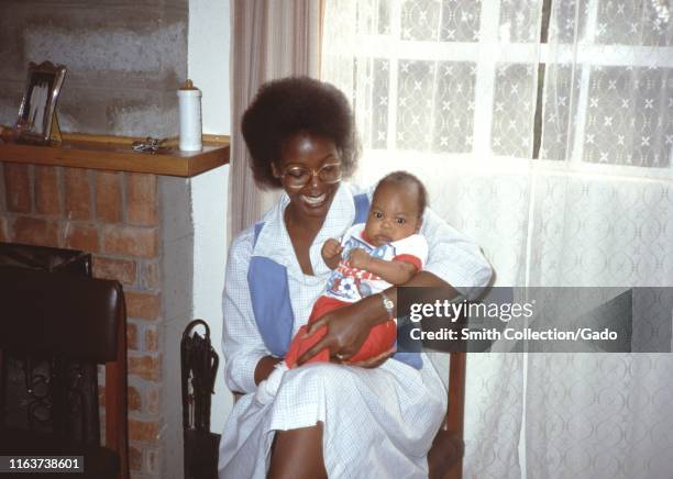 African-American mother smiling and holding child while sitting in a chair in a domestic living room, 1965.