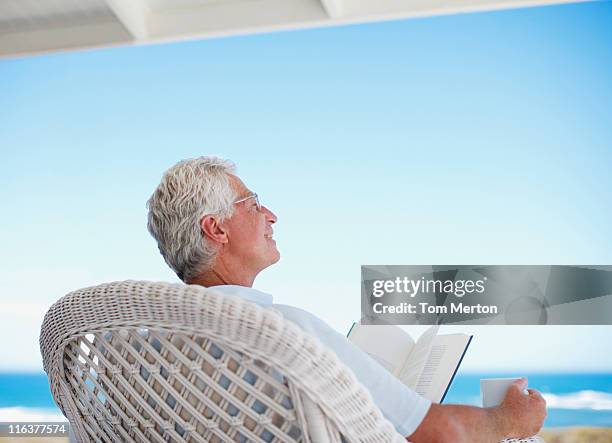 homme âgé lisant un livre sur la plage terrasse - chaise de dos photos et images de collection