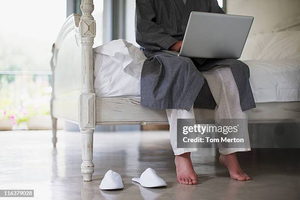 senior man with bare feet sitting on edge of bed using laptop - old man feet stock pictures, royalty-free photos & images
