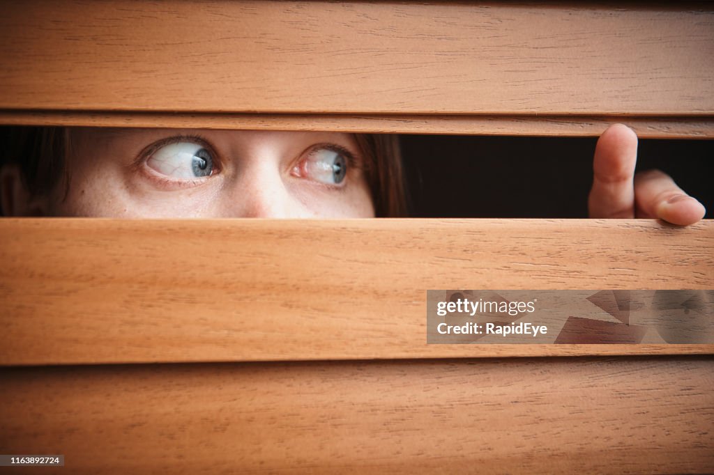 Terrified young woman peeks sideways through wooden venetian blinds