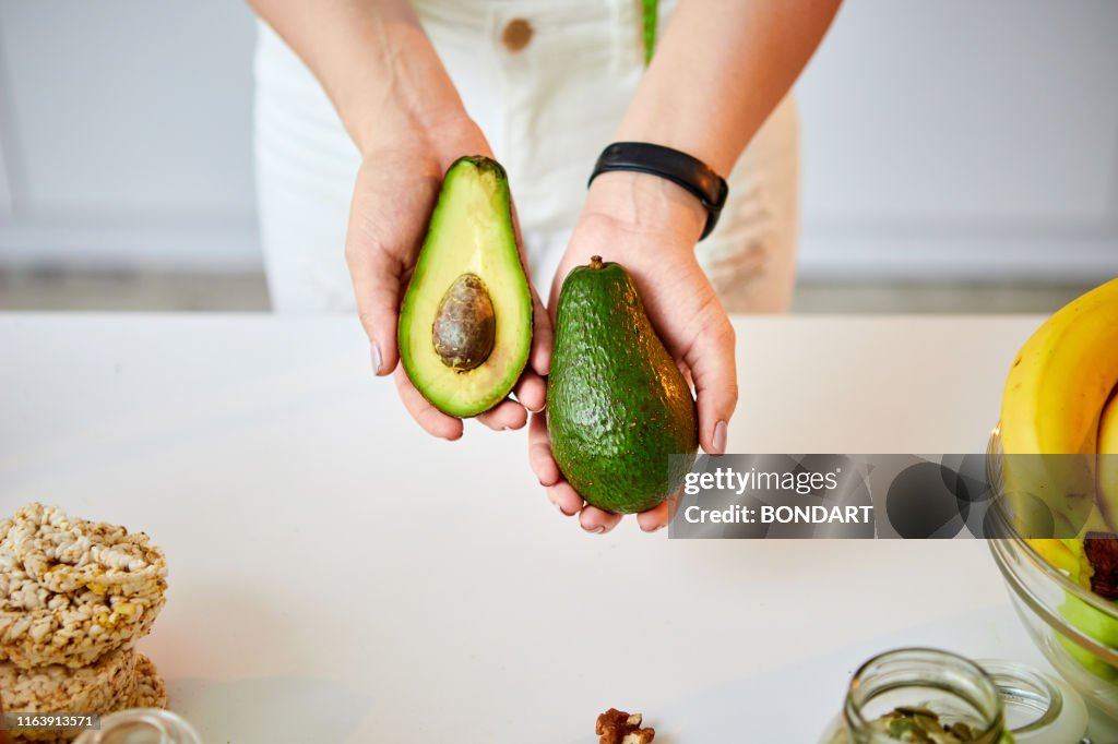 Young happy woman holding avocado for making salad in the beautiful kitchen with green fresh ingredients indoors. Healthy food and Dieting concept. Loosing Weight