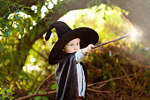 Little boy in pointed hat and black cloak playing with magic wand outdoors. Little wizard.