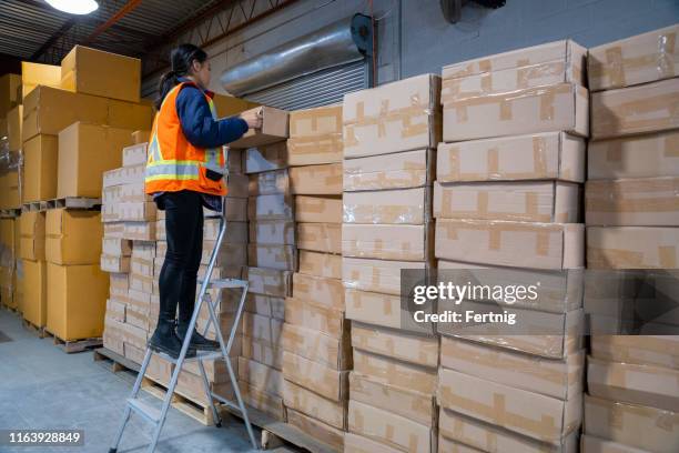 an industrial warehouse workplace safety topic. a female employee uses a short step ladder to reach some higher placed merchandise. - step stool stock pictures, royalty-free photos & images